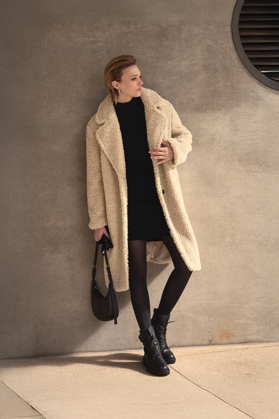 Γυναίκα με κρεμ παλτό teddy, μαύρο φόρεμα και μαύρη τσάντα, κοιτάζει δεξιά.