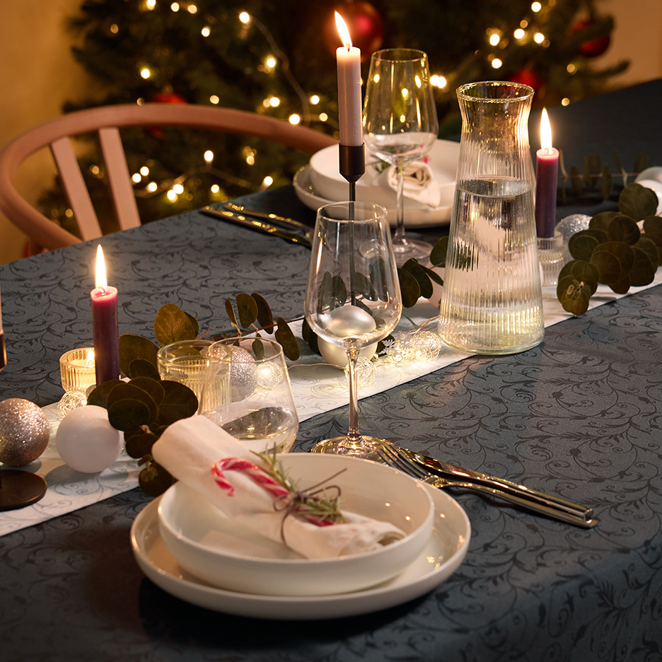 Χριστουγεννιάτικο τραπέζι με κεριά, γυάλινα σκεύη και λευκά σερβίτσια σε ένα τραπεζομάντιλο με σχέδια.