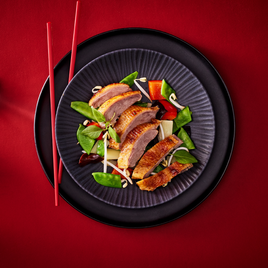 Φέτες τραγανής πάπιας με σοταρισμένα λαχανικά, φύτρες φασολιών και βασιλικό σε μαύρο πιάτο.