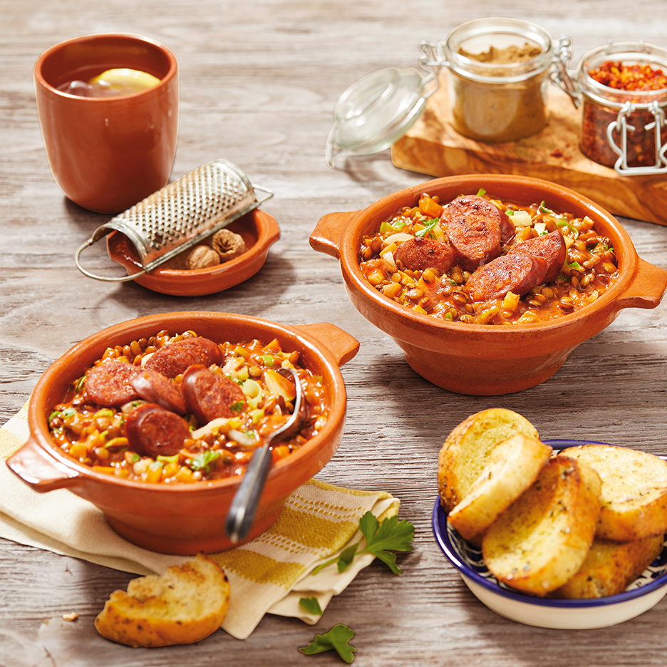 Δύο μπολ φακές με λουκάνικο, σκορδόψωμο και ένα ποτό σε ξύλινο τραπέζι.