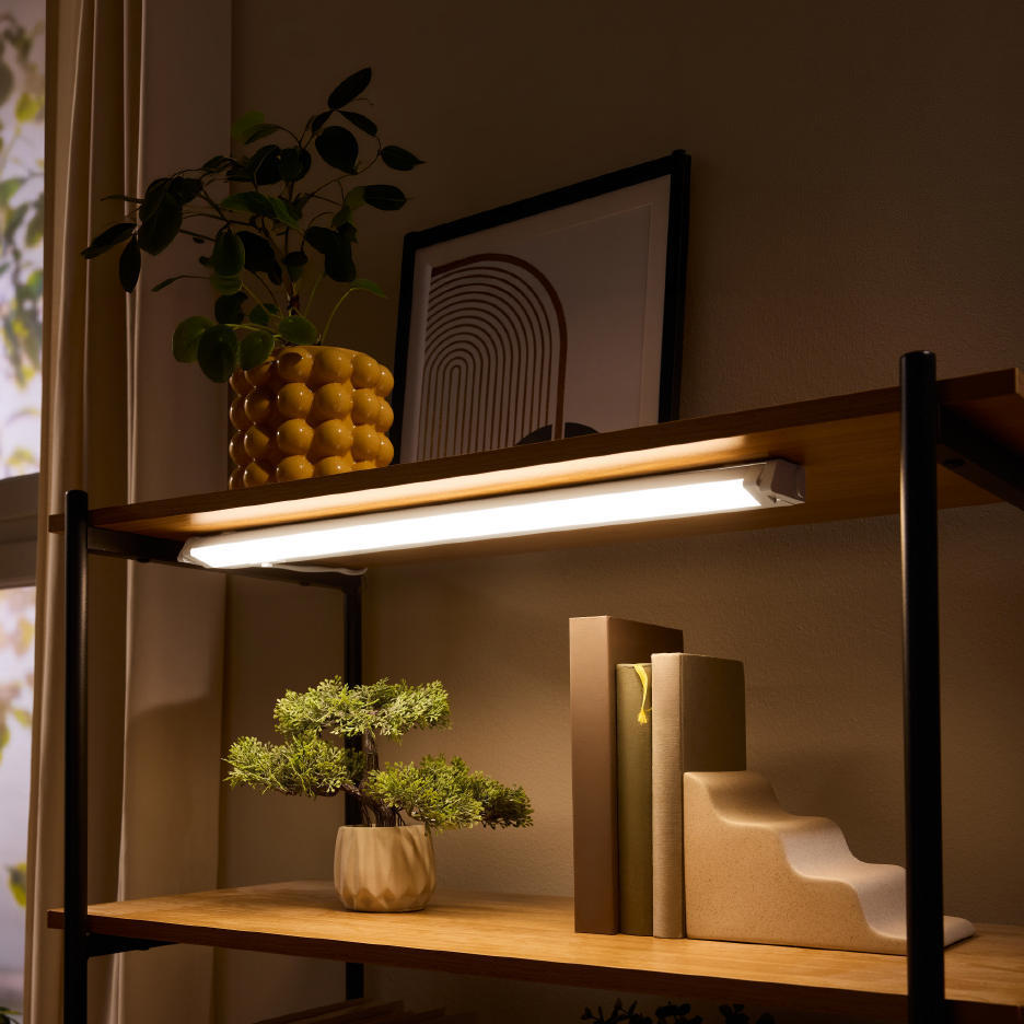 Ράφι βιβλιοθήκης με φωτιστικό κάτω από το ράφι, φυτό σε κίτρινη γλάστρα και διακοσμητικά.
