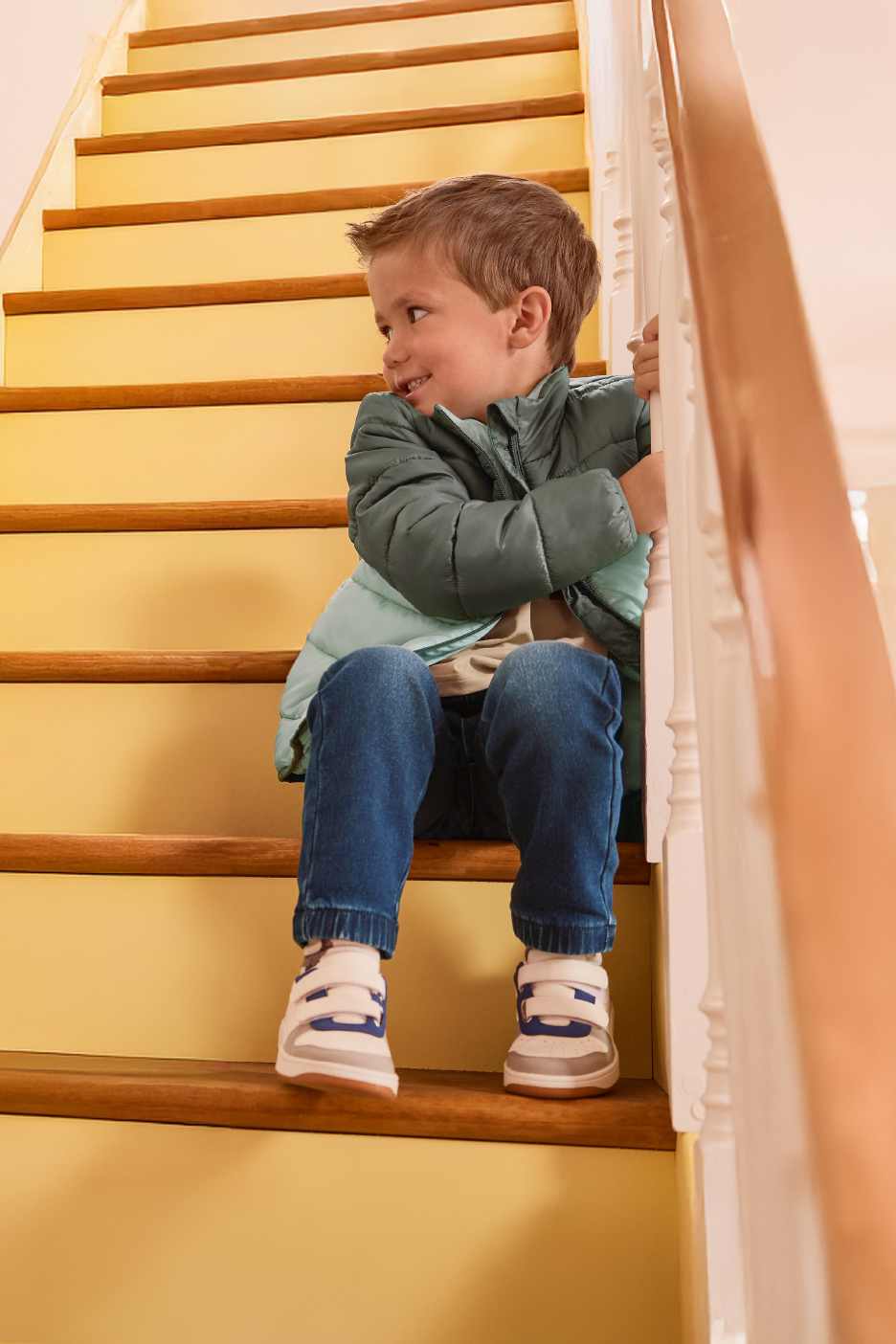 Χαμογελαστό αγόρι με πράσινο φουσκωτό μπουφάν, μπλε τζιν και αθλητικά παπούτσια κάθεται σε ξύλινη σκάλα.