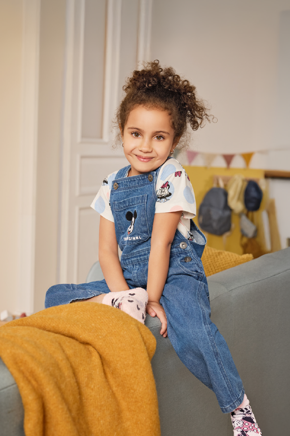 Χαμογελαστή κοπέλα με τζιν ολόσωμη φόρμα και μπλουζάκι με σχέδια, κάθεται σε καναπέ.