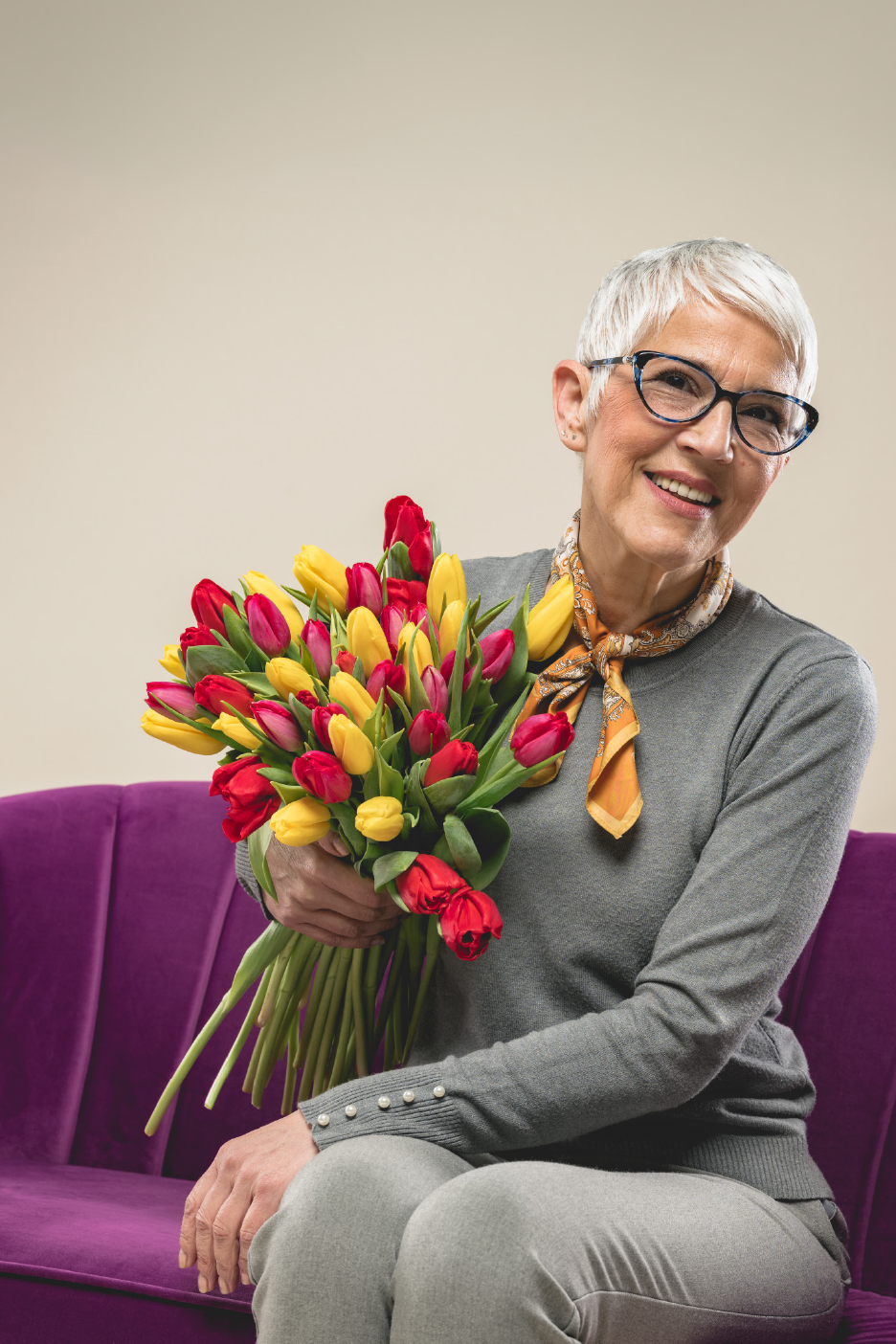 Χαμογελαστή ηλικιωμένη γυναίκα κρατάει μια μεγάλη ανθοδέσμη με κόκκινες και κίτρινες τουλίπες.