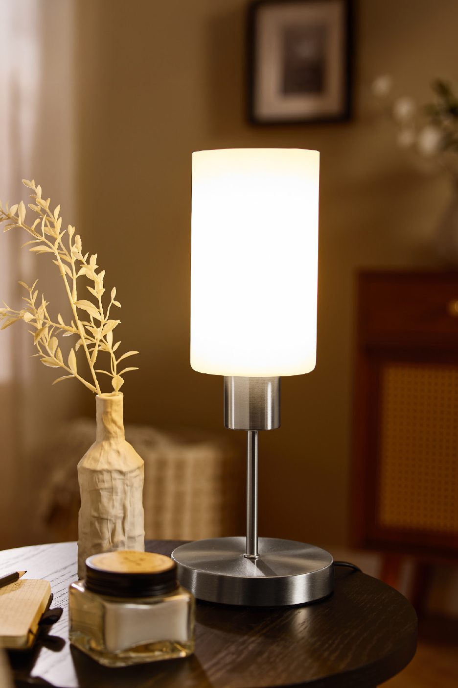 Μια μοντέρνα επιτραπέζια λάμπα με λευκό κυλινδρικό αμπαζούρ και βουρτσισμένη μεταλλική βάση, δίπλα σε ένα διακοσμητικό βάζο.