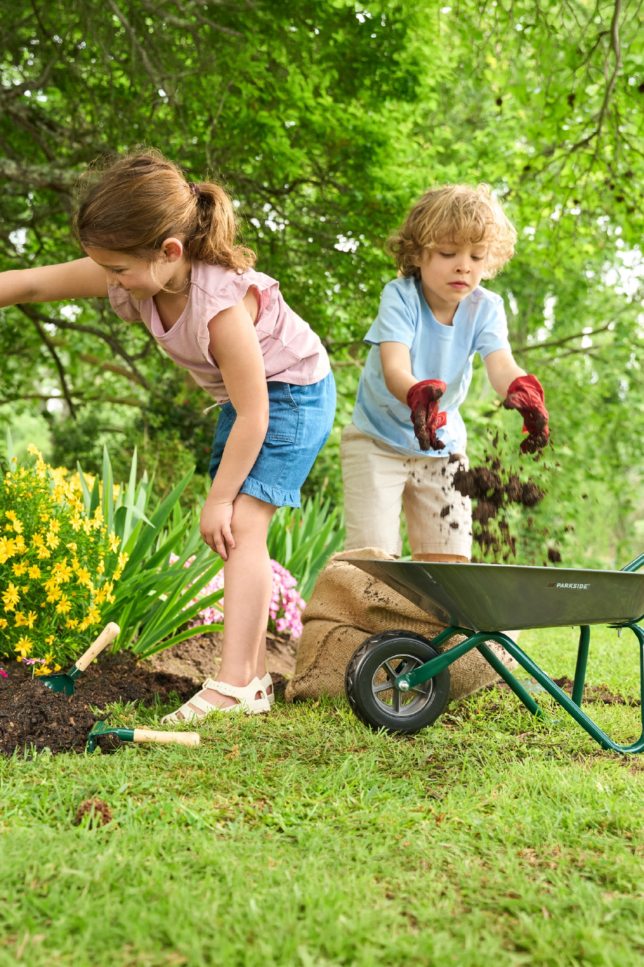 Δύο παιδιά κηπουρεύουν, το ένα βάζει χώμα σε καρότσι, με εργαλεία κηπουρικής κοντά.