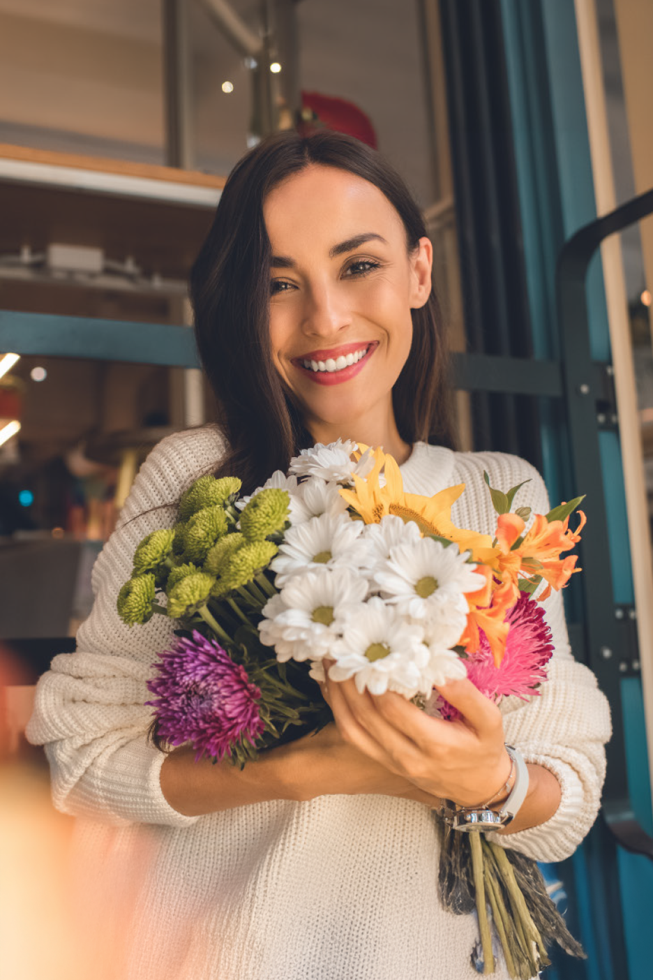 Χαμογελαστή γυναίκα με λευκό πουλόβερ κρατάει ένα ζωντανό μπουκέτο φρέσκα λουλούδια.