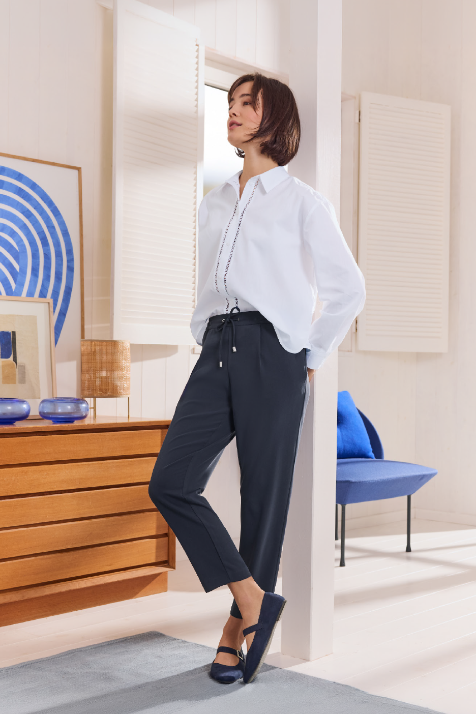 Γυναίκα με λευκό πουκάμισο και σκούρο μπλε παντελόνι, ακουμπισμένη σε τοίχο σε φωτεινό δωμάτιο.