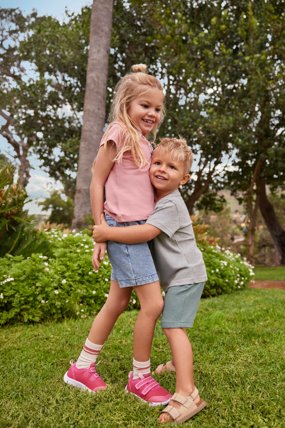 Δύο χαρούμενα παιδιά, ένα κορίτσι με ροζ μπλουζάκι και τζιν σορτς, και ένα αγόρι με γκρι μπλουζάκι και πράσινο σορτς, αγκαλιάζονται σε εξωτερικό χώρο.