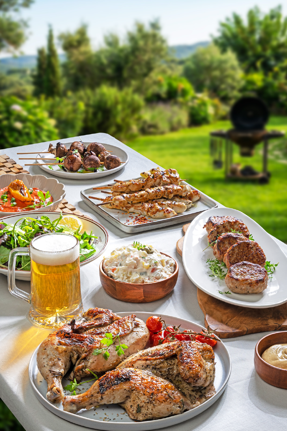 Ένα τραπέζι γεμάτο με ψητό κοτόπουλο, σουβλάκια, μπιφτέκια, σαλάτες και μπύρα σε έναν κήπο.