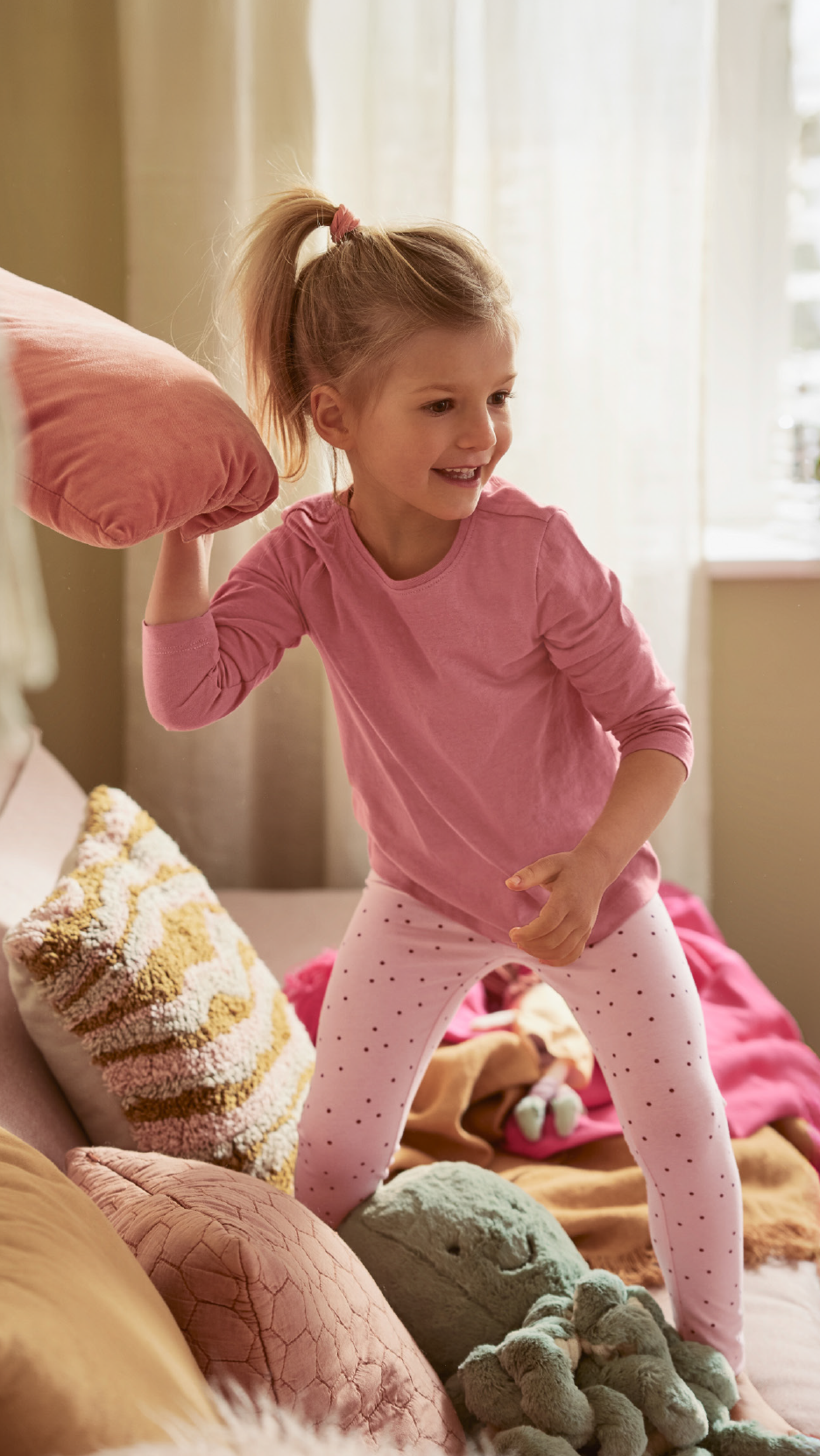 Χαμογελαστή κοπέλα με ροζ πιτζάμες παίζει με ένα ροζ μαξιλάρι σε κρεβάτι με μαξιλάρια.