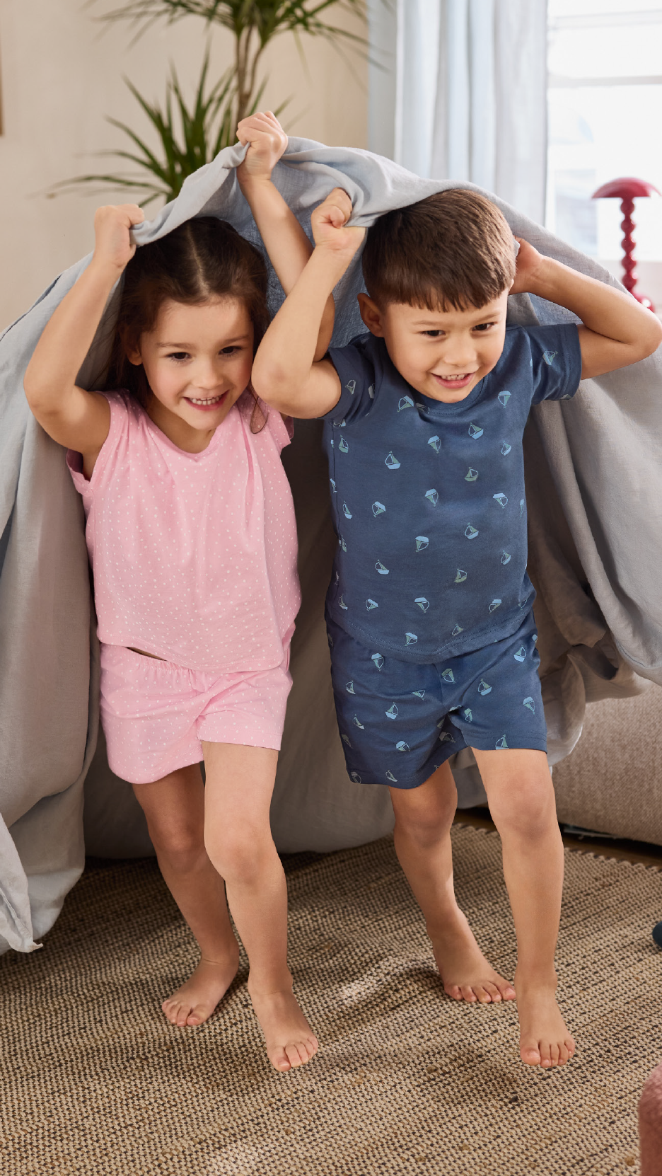 Δύο χαρούμενα παιδιά με κοντές πιτζάμες παίζουν κάτω από μια κουβέρτα.