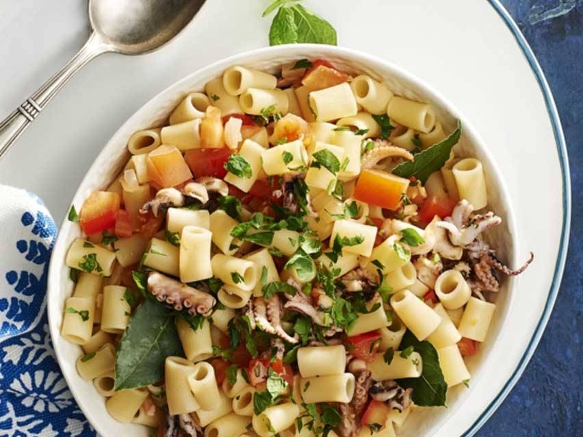 Σαλάτα ζυμαρικών με χταπόδι, ντομάτες και μυρωδικά σε ένα λευκό μπολ.